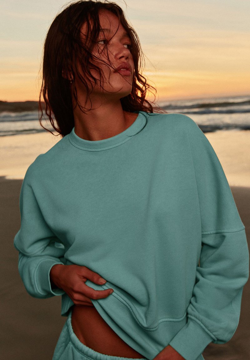 Jeune femme aux cheveux mouillés portant un sweat large bleu sarcelle, debout sur une plage au coucher du soleil, regardant sur le côté tout en soulevant son sweat-shirt de la main.