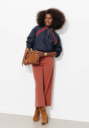 Femme avec coiffure afro portant une veste bleu marine, un pantalon couleur rouille, des bottes en daim beige, tenant un sac en daim beige assorti, appuyée contre un mur blanc.