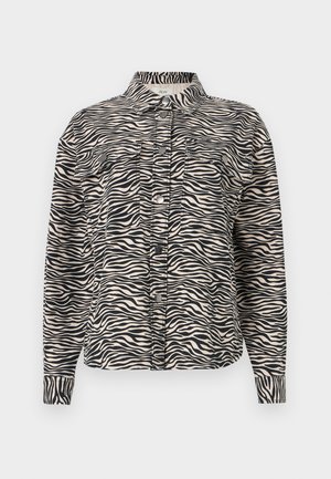 Long-sleeve button-up shirt in beige with black zebra stripes, featuring a collar, two chest pockets, and black snap buttons.