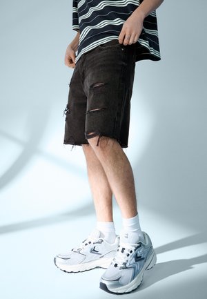 Bărbat purtând pantaloni scurți negri rupți, șosete albe, pantofi sport albi și gri și o cămașă în dungi alb-negru, stând în picioare pe un fundal simplu.