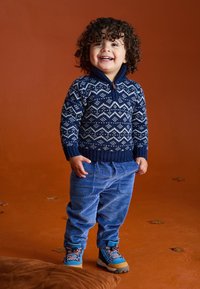 Marineblauwe trui met wit zigzagpatroon, geribbelde manchetten en een halve rits; blauwe corduroy broek; veelkleurige wandelschoenen; oranje achtergrond.