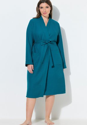 Frau, die barfuß steht und einen türkisfarbenen, strukturierten Bademantel trägt, der in der Taille gebunden ist, lange Ärmel und zwei vordere Taschen hat.