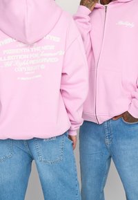 Haut zippé rose en tissu doux, avec des graphiques textuels blancs au dos et un logo brodé sur le côté gauche de la poitrine.