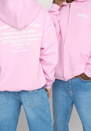 Haut zippé rose en tissu doux, avec des graphiques textuels blancs au dos et un logo brodé sur le côté gauche de la poitrine.