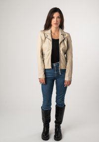 Beige leren motorjack met ritsen over een zwarte top, gecombineerd met blauwe jeans en kniehoge zwarte laarzen. Gladde textuur, getailleerd ontwerp.