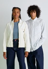 En kremfarget zip-jakke over en lys blå topp, sammen med mørk denim. Den mannlige modellen har på seg en hvit skjorte med blå striper.