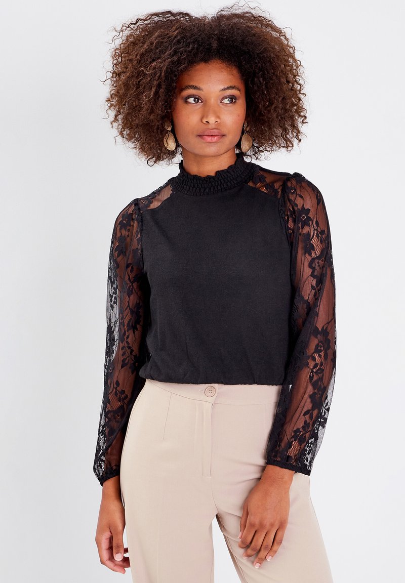 Haut noir avec des manches longues en dentelle, encolure smockée texturée et coupe ajustée. Porté avec un pantalon beige ; détails délicats en dentelle florale sur les manches.