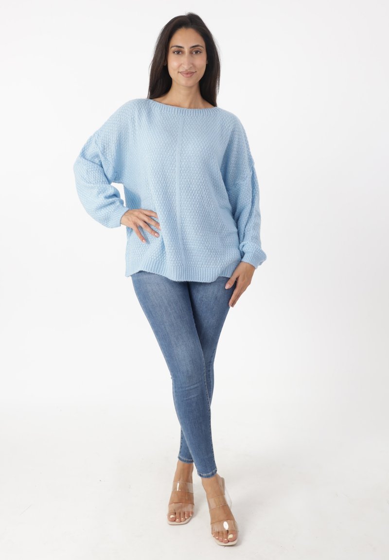 Suéter de punto azul claro con un patrón texturizado, amplio escote barco y hombros caídos, combinado con unos jeans de mezclilla ajustados y sandalias transparentes.