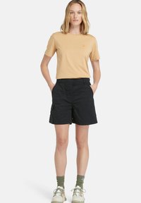 T-shirt di cotone beige chiaro con scollo rotondo, abbinato a pantaloni corti neri fino al ginocchio con tasche laterali. Scarpe da ginnastica bianche e calzini verdi completano l'outfit.