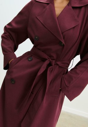 Femme portant un trench-coat bordeaux à double boutonnage avec des revers larges et une ceinture nouée, les mains dans les poches.