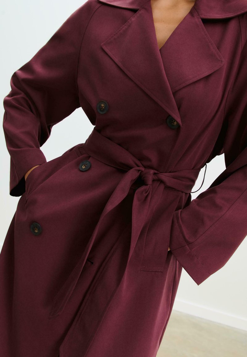 Nainen, jolla on yllään kaksirivinen, syvän burgundinvärinen trench-coat, jossa on leveät kaulukset ja vyötärölle sidottu vyö, kädet taskuissa.