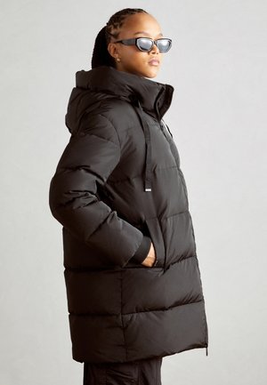 Doudoune noire avec capuche, présentant un design matelassé, des poignets élastiques et des cordons de serrage au niveau du cou. Texture lisse avec une coupe décontractée.