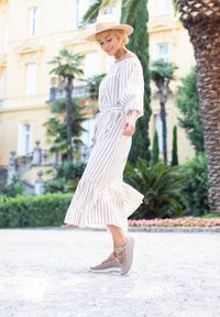 Gestreiftes, schulterfreies, langes Kleid mit einem Rüschensaum in Creme und Beige, kombiniert mit hellbeigen Schnürsneakern auf einem Kiesweg.