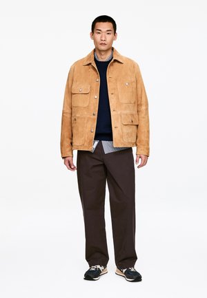 Brązowa zamszowa kurtka z czterema przednimi kieszeniami, zapięciem na guziki i kołnierzem z klapą, noszona na granatowym swetrze i koszuli w paski, zestawiona z ciemnymi spodniami.