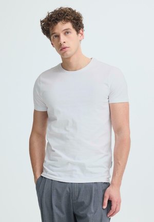 Młody mężczyzna o kręconych włosach, ubrany w gładki biały T-shirt z krótkim rękawem i szare spodnie, stojący na jasnym tle.