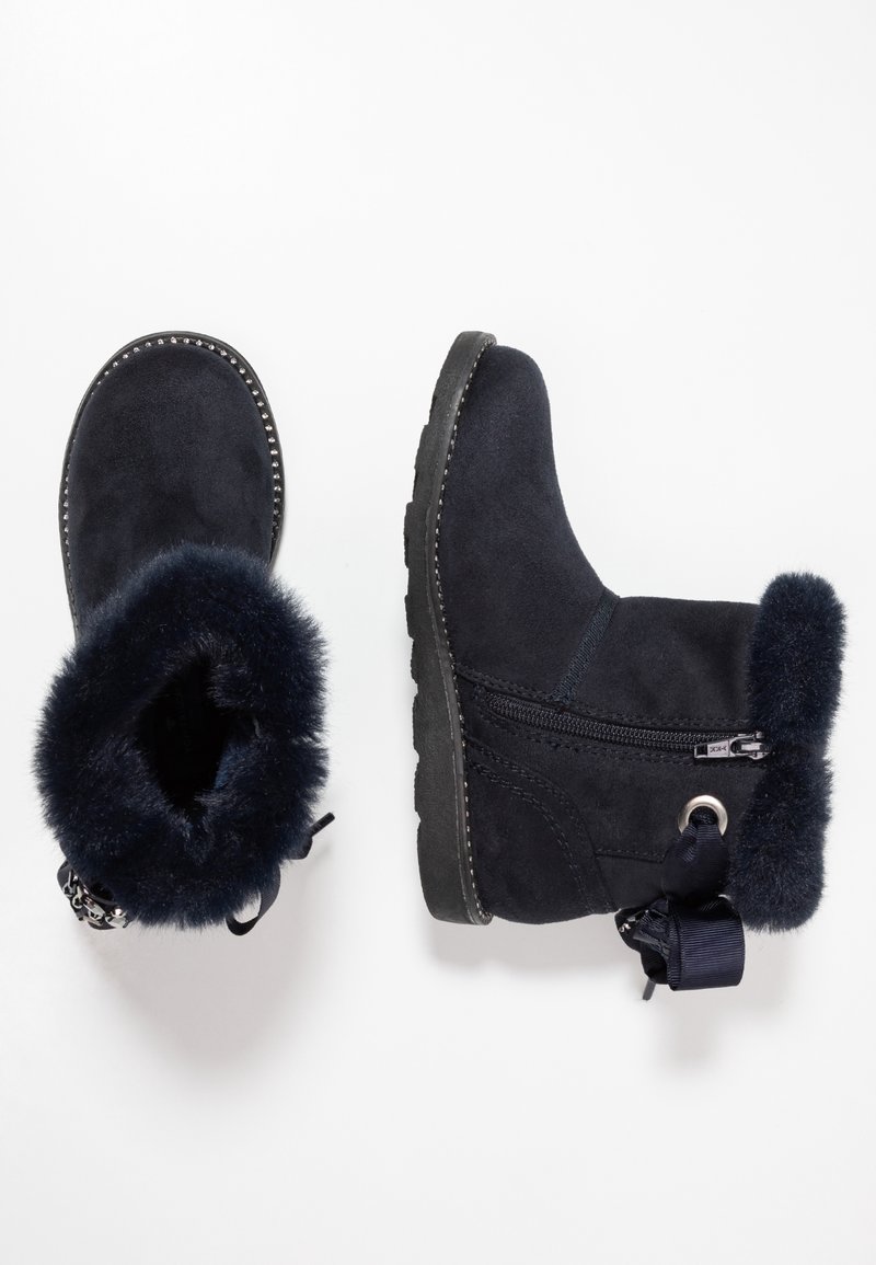 Paire de bottes d’hiver noires avec doublure en fourrure, fermeture éclair latérale, nœud décoratif et bordure cloutée sur la semelle sur fond blanc.