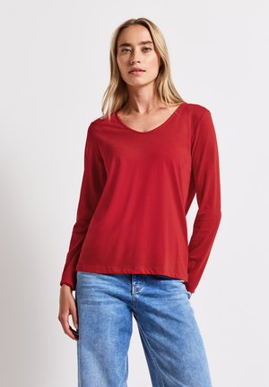 T-shirt rouge à manches longues avec un col en V, fabriqué à partir d'un tissu lisse ; associé à un jean bleu clair.