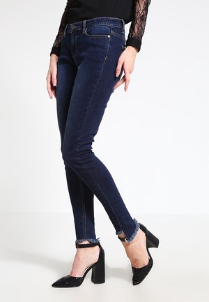 Kobieta nosząca ciemnoniebieskie dopasowane dżinsy z postrzępionymi brzegami i czarne buty na wysokim obcasie z paskami wokół kostek, stojąca bokiem.