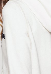 Krémová mikina s měkkou texturou, z bočního pohledu s viditelnými švy a kapucí. Černý textový detail na přední straně.