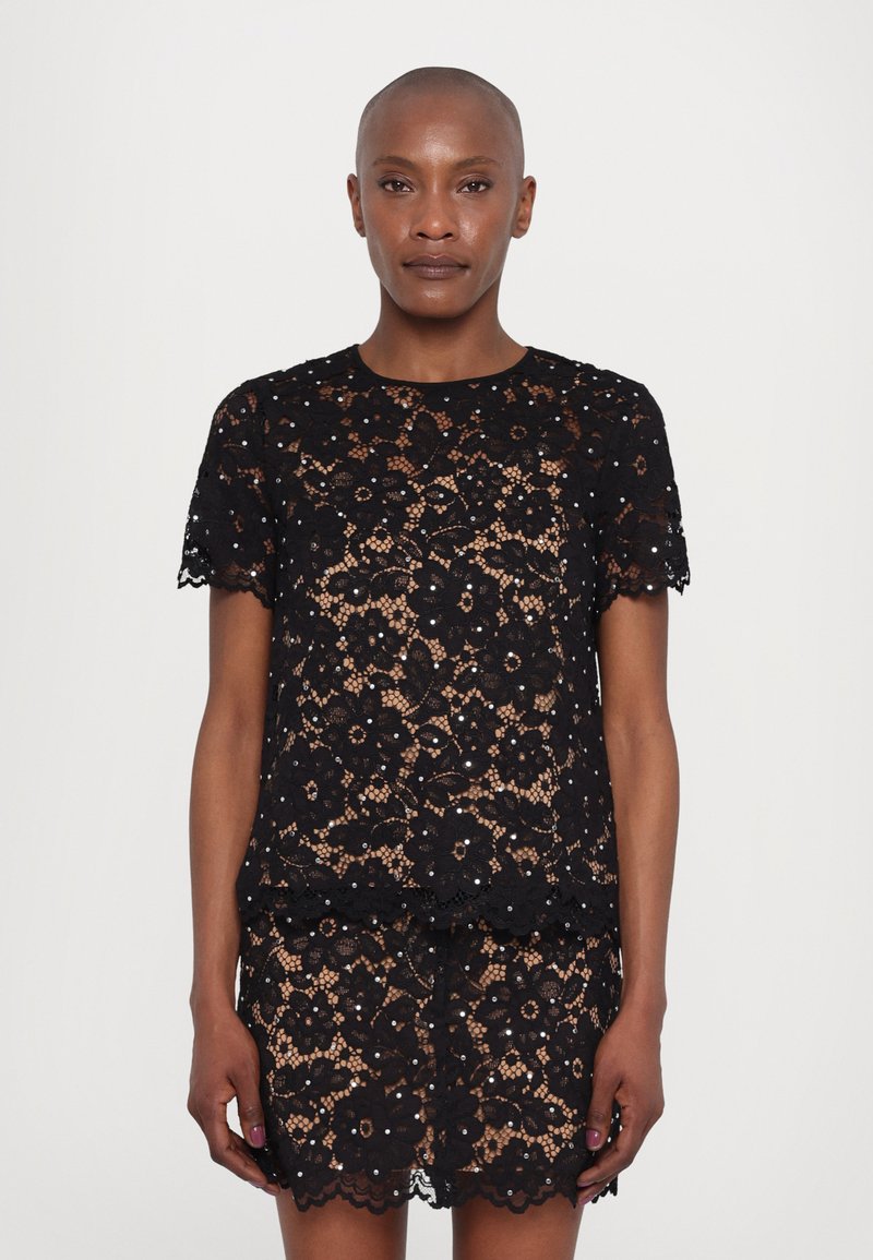 Top en dentelle noire avec motif floral, manches courtes, bords festonnés, superposé sur une jupe en dentelle noire coordonnée. Tissu texturé et semi-transparent.