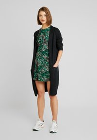 Svart strikket cardigan over en grønn blomstret kjole. Cardiganen har lange ermer og lommer; kjolen har et tettsittende liv og vid skjørt.