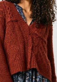 Rostfarbener gestrickter Pullover mit V-Ausschnitt, der ein strukturiertes Muster, lange Ärmel und ein kurzes Design aufweist, das über einem blümigen Kleid getragen wird.