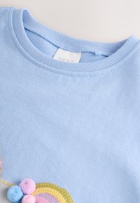 T-shirt en coton bleu clair avec un col rond et des accents multicolores de pompons en rose, bleu et jaune près de l'ourlet.