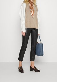 Gilet en tricot beige sur une chemise blanche, associé à un pantalon en cuir noir, des mocassins marron et un sac à main bleu. Foulard en soie marron à motifs noué autour du cou.