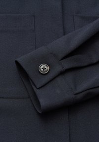 Primo piano di tessuto blu marina scuro che mostra il polsino di una manica di camicia con un singolo bottone nero a quattro fori e bordi della tasca cuciti.