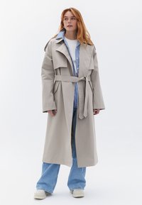 Beige Trenchcoat mit breiten Revers, Taillengürtel und langen Ärmeln, über einem blau-weißen gestreiften Hemd getragen. Styliert mit lockeren Jeans.