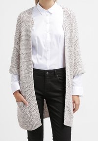 Stickad cardigan i ljusbeige med öppen front, korta ärmar och strukturerat mönster. Bärs över en vit skjorta och svarta byxor.