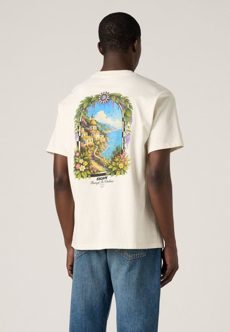 Man wearing white t-shirt with colorful seaside village and floral arch graphic and text "Escape Through the Window" on the back.