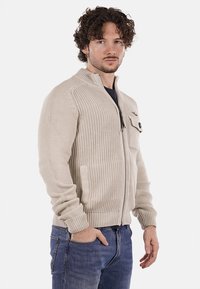 Cardigan in maglia beige con zip e colletto alto, tessuto texturizzato e tasca a toppa. Design aderente con polsini e orlo a costine.