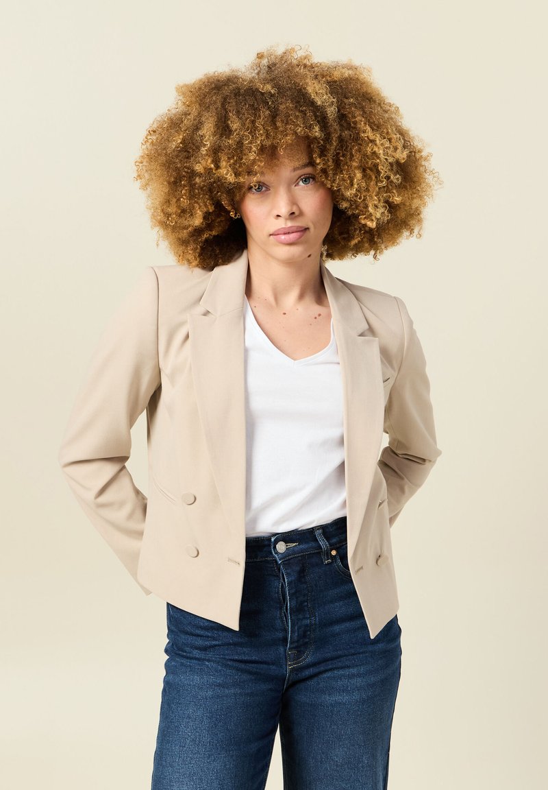 Blazer beige doppiopetto con revers a punta, realizzato in tessuto liscio. Indossato sopra una camicia bianca a V, abbinato a jeans in denim scuro.