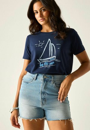 Kobieta w granatowym t-shircie z białym żaglówką i napisem „by the sea” w połączeniu z jasnoniebieskimi, wysokim stanem szortami jeansowymi.