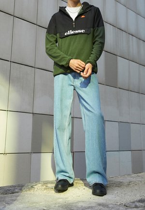 Person trägt einen grünen und schwarzen ellesse-Pullover, hellblaue Jeans, schwarze Schuhe, steht vor einer grauen gefliesten Wand.