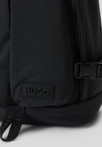 Mochila preta de nylon com uma superfície texturizada, bolso com fecho e uma etiqueta retangular com o logo "HUGO" em relevo. Inclui tira ajustável.