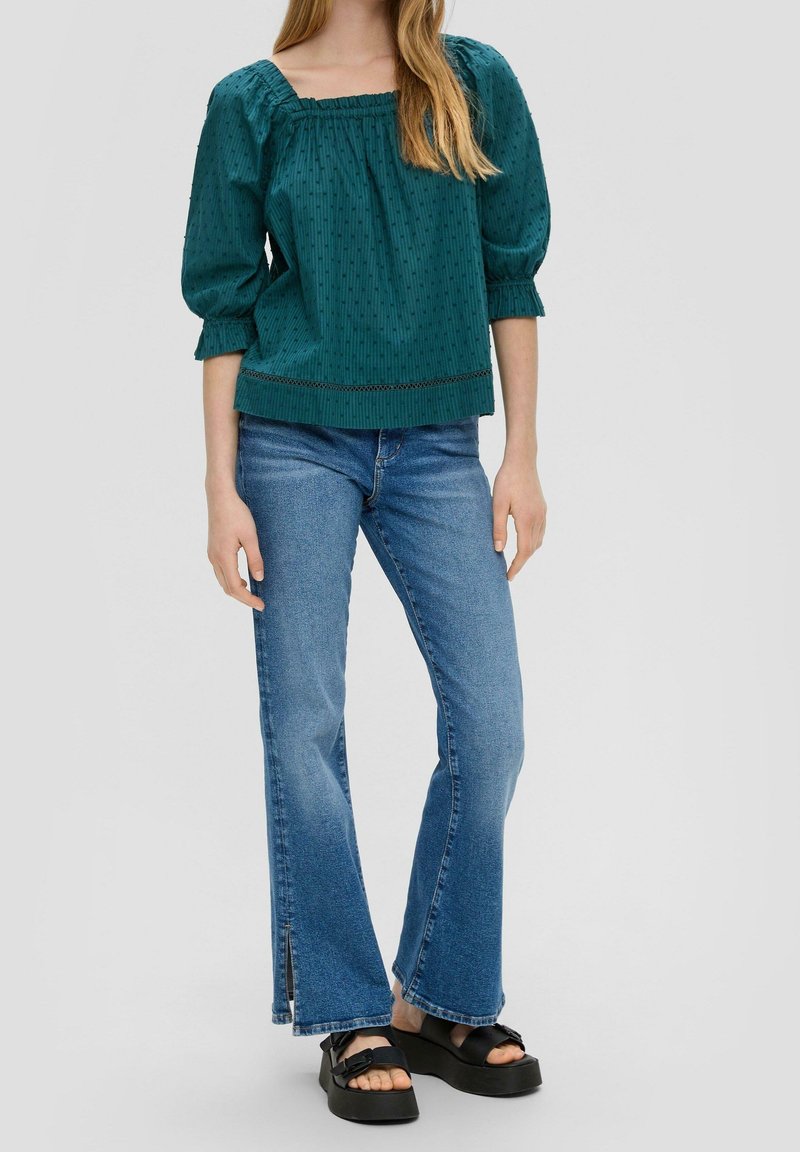 Kobieta w turkusowej bluzce o fakturze z rękawami 3/4, niebieskich dżinsach z wysokim stanem i rozkloszowanym krojem, z przednimi rozporkami, oraz czarnych sandałach na platformie.