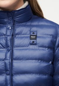 Κοντινό πλάνο ενός ατόμου που φοράει μπλε καπιτονέ μπουφάν με κουμπιά πάτωσης και την ετικέτα "Blauer U.S.A." στο στήθος.