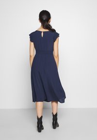 Nainen, jolla on tummat hiukset poninhännällä, pukeutuu tummansiniseen midi-mekkoon, jossa on lepattelevat hihat, ja mustiin nilkkapolkimiin, seisoa tasaisella taustalla.