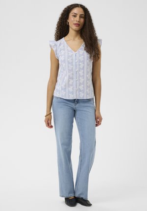 Femme aux cheveux bouclés portant un haut sans manches bleu clair à motif floral et un jean bleu clair, debout devant un fond uni.