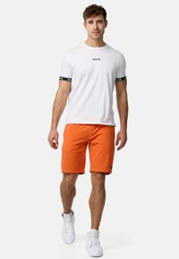 Hvid t-shirt med korte ærmer og sorte accentstriber, parret med orange shorts. Model iført hvide atletiske sneakers.