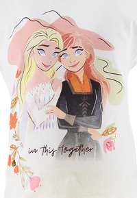 Camisa blanca con personajes al estilo de dibujos animados que representan a dos mujeres con cabello rubio y castaño rojizo. Diseño floral y texto: "juntas en esto."