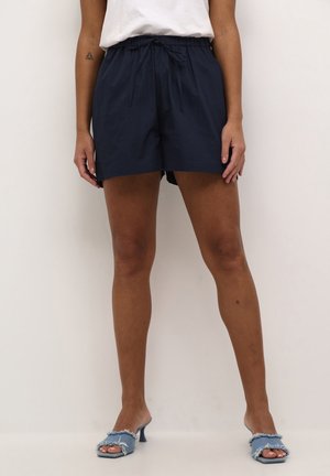 Person iført marineblå shorts med snøre, en hvid top og lyseblå denim sandaletter med hæl, stående mod en ensartet baggrund.
