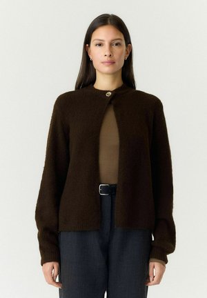 Vrouw met lang donker haar die een donkergbruine zachte cardigan draagt, dichtgeknoopt bij de hals, een bruin topje en donkere broek met een zwarte riem.