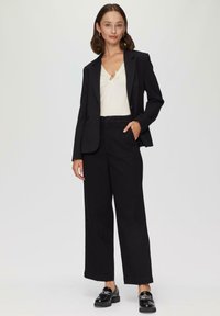 Zwarte op maat gemaakte blazer over een witte top met een knoopdetail, gecombineerd met zwarte wijde broek en glanzende zwarte loafers.
