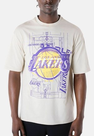 Homme portant un t-shirt beige avec plusieurs logos superposés des Los Angeles Lakers et un diagramme de terrain de basket en violet et jaune.