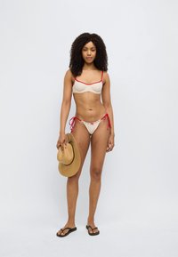 Σετ μπικίνι με κρεμ τοπ με κόκκινες λεπτομέρειες και πουά σχέδιο. Παντελόνια με κορδόνια στο πλάι. Το μοντέλο κρατάει ένα καπέλο από ψάθα σε ταμπά χρώμα, φορώντας μαύρες σαγιονάρες.