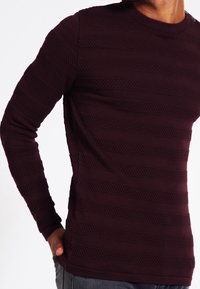 Burgundowy sweter z długim rękawem, wykonany z dzianiny, z teksturowanymi poziomymi paskami i okrągłym dekoltem, o dopasowanym kroju i ściągaczami przy rękawach.
