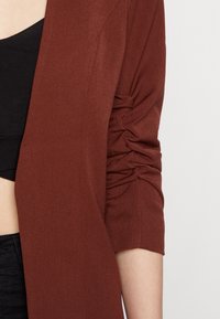 Svetrový cardigan v barvě rezavé mědi z měkké strukturované tkaniny s nařaseným detailem na loktech. Otevřený přední design sahá ke třičtvrtinovým rukávům.